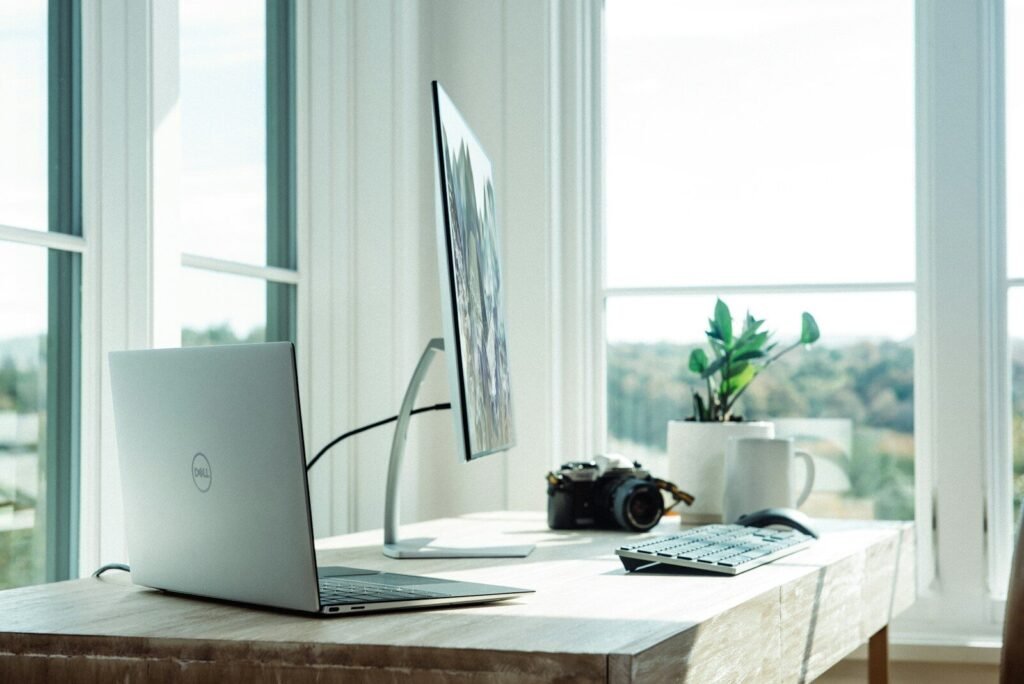 Laptop, monitor, webcam, and plant on a wooden desk in a sunlit home office, illustrating home office gadgets for remote workers